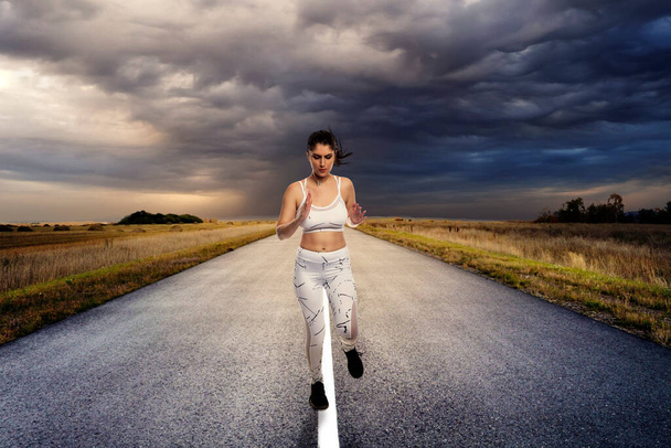 Pasująca kobieta ubrana w odzież sportową biegająca lub biegająca samotnie na odosobnionej drodze do ćwiczeń podczas dystansu społecznego. Jest zdeterminowana, by utrzymać zdrowy tryb życia. - Zdjęcie, obraz