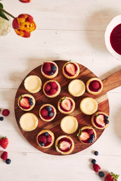 A high angle shot of cheese cupcakes with fruit jelly and fruits on a wooden plate with a white background - Фото, зображення