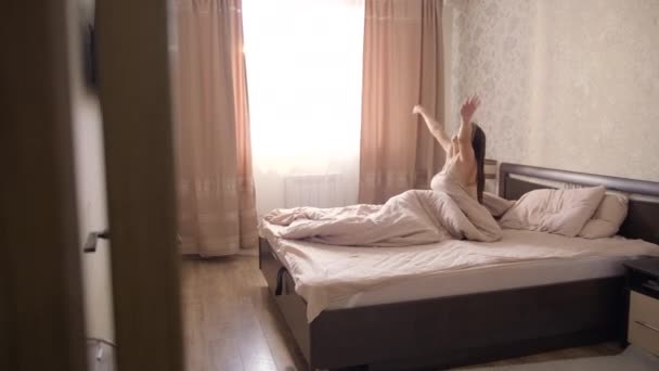 A young girl with long dark hair in a light nightgown wakes up in a large bed in the bedroom room and opens the curtains on the windows - Materiaali, video