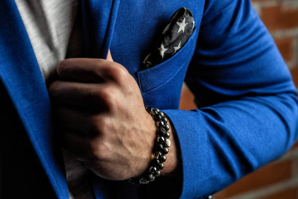 Браслеты талисманы драгоценных камней и минералов на руке, в синем деловом костюме на фоне кирпичной стены
 - Фото, изображение