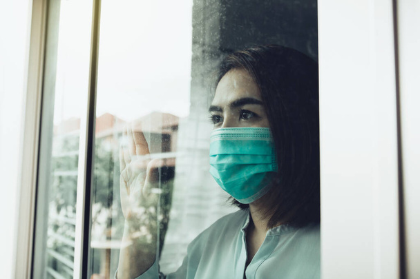Asian woman looking through the window and wearing medical mask protection from the illness with stay isolated quarantine COVID-19 pandemic virus mask against coronavirus disease at home. - Foto, Imagem