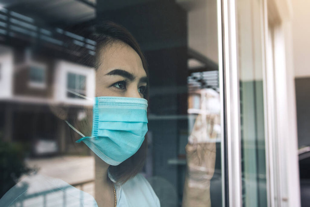 Asian woman looking through the window and wearing medical mask protection from the illness with stay isolated quarantine COVID-19 pandemic virus mask against coronavirus disease at home. - Zdjęcie, obraz