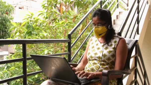 Primer plano de una mujer india con máscara de seguridad y micrófono de auriculares, trabajando con un portátil sentado en el balcón durante la pandemia del virus de la corona Covid19
 - Imágenes, Vídeo