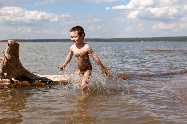 Ein fröhlicher kleiner Junge genießt den Sommer und spielt an einem warmen Sommertag im Wasser, Spritzer aus dem Meer fliegen in verschiedene Richtungen, im Hintergrund Strand und Wald - Foto, Bild
