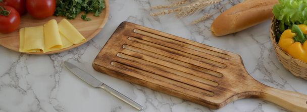 Кухонный стол из мрамора с деревянным подносом, сыром, овощами, французским багетом и столовыми приборами
  - Фото, изображение