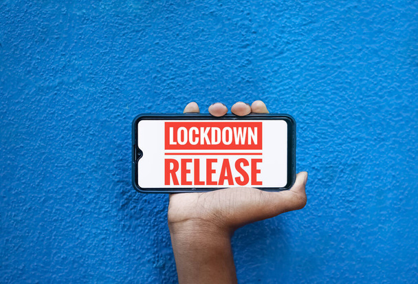 Lock down release wording on smart phone screen isolated on blue background with copy space for text. Person holding mobile on his hand and showing front screen. Lock down release for corona virus . - Photo, Image