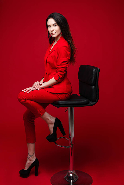 Charmante blanke vrouw met lang donker recht haar in rood kantoorpak, zwarte schoenen zit op zwarte stoel aand poseert voor de camera - Foto, afbeelding