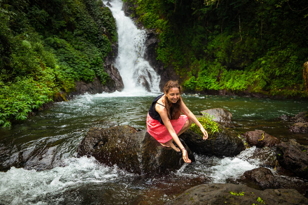 若い白人女性が岩の上に座って、滝の近くの川で水で遊んでいます。旅行のコンセプト。黒とピンクのドレスを着た女性。インドネシアバリのサンバンガンにあるデダリの滝 - 写真・画像