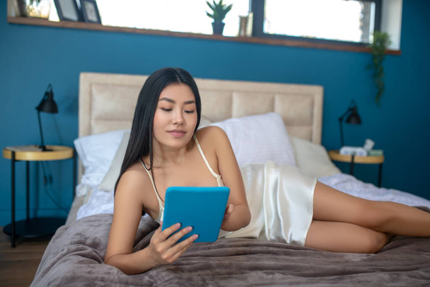 Young brunette enjoying her morning, lying on her side, looking at tablet - Photo, Image