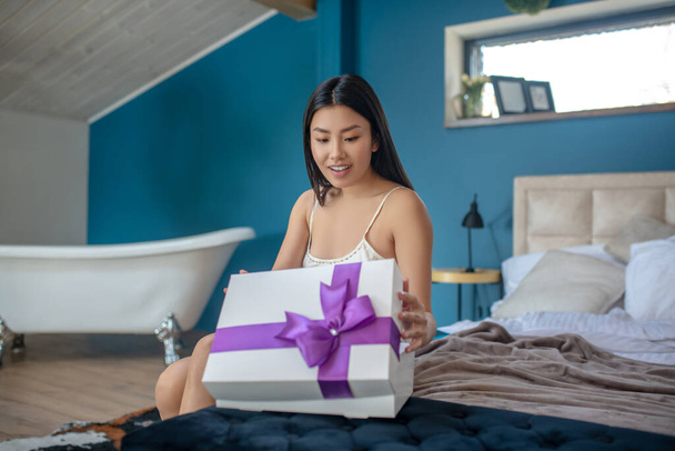 Young brunette in night dress sitting on bed, smiling, opening present box with purple ribbon, smiling - 写真・画像