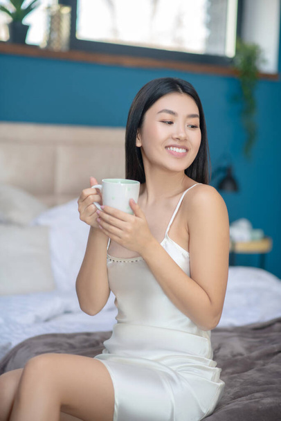 Young brunette in night dress sitting on bed, holding white cup, drinking, smiling - Photo, image