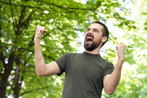 Бородатий молодий кавказький чоловік, радісний, проходить через парк між деревами, піднімаючи руки вгору. Емоційне вираження радості
 - Фото, зображення
