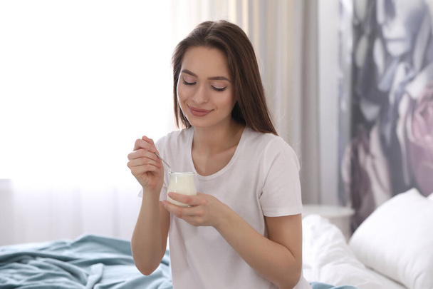 Young attractive woman with tasty yogurt on bed at home - Photo, Image