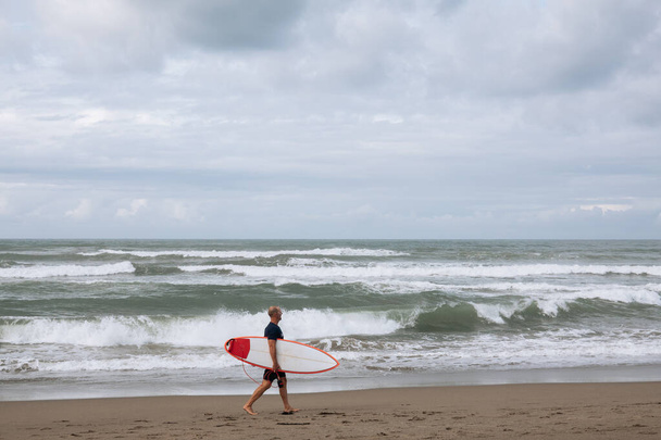 CANGGU, BALI, INDONESIA - FEBRUARI 25, 2019: jonge surfer man wandelen op het strand met surfplank in de zomer aan de kust van Bali eiland. Recreatie en watersport tijdens vakantie in Indonesië - Foto, afbeelding