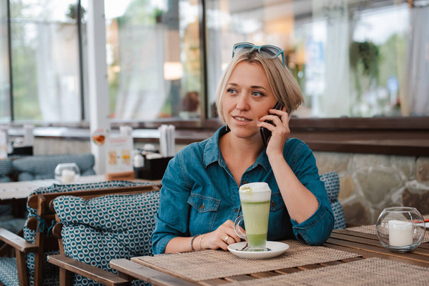 Młoda kobieta pije ciesząc się matcha zielona herbata latte w letni poranek w kawiarni rozmowy przez telefon komórkowy smartphone. Blondynka jest młodą damą z koncepcją zdrowego odżywiania. - Zdjęcie, obraz