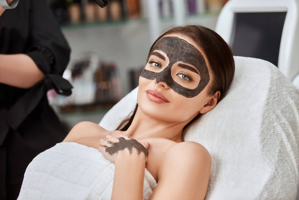 顔に炭素マスクをした可愛い女性がスパに横になってカメラや美容方法や顔の治療を探しています - 写真・画像