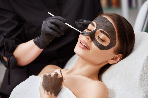 close-up van vrouw gezicht liggen in salon en schoonheidsspecialiste toepassen van carbon masker, mooi meisje met huidverzorging procedures in spa - Foto, afbeelding