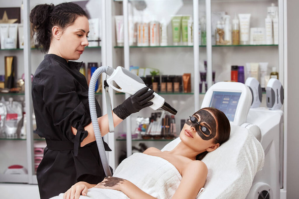 αισθητικός κρατώντας μηχανή κοντά στο πρόσωπο γυναίκα που καλύπτεται με μάσκα άνθρακα στο σαλόνι σπα, όμορφο κορίτσι λαμβάνει θεραπεία προσώπου με λέιζερ στην κλινική ομορφιάς - Φωτογραφία, εικόνα