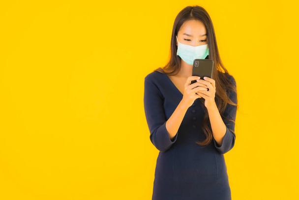 Portré gyönyörű fiatal ázsiai nő maszk és okos mobiltelefon vagy mobil sárga elszigetelt háttér - Fotó, kép