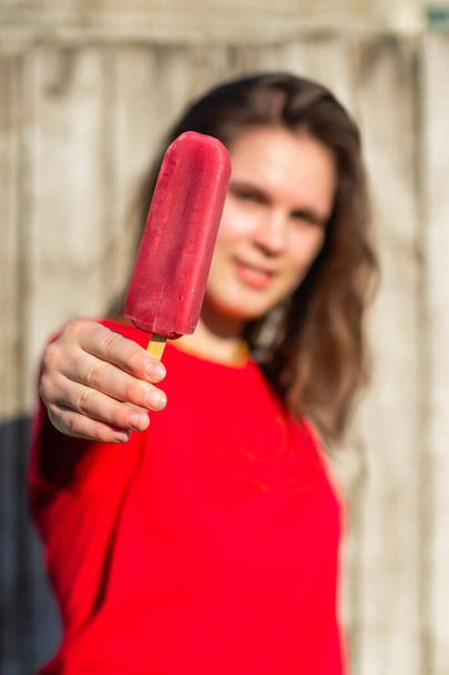 Teenage girl with pink ice cream outdoors. Young brunette woman in red t shirt against wooden wall fence eating delicious cream. Concept of dessert, carefree, joy, teenage lifestyle, fun, emotions. - Photo, image