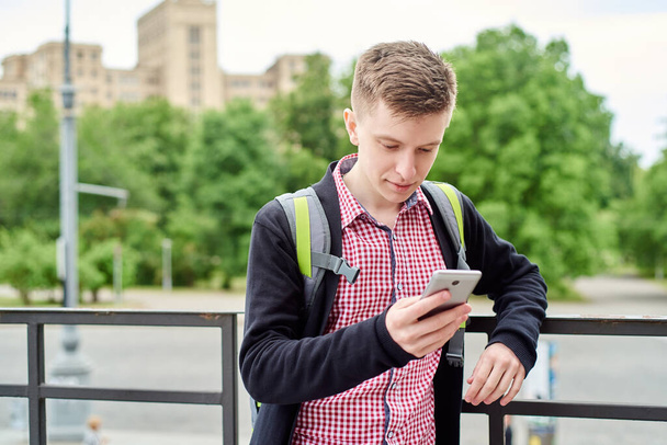 Sırt çantalı mutlu genç bir öğrencinin portresi parkta yürüyor ve açık havada cep telefonu kullanıyor. - Fotoğraf, Görsel