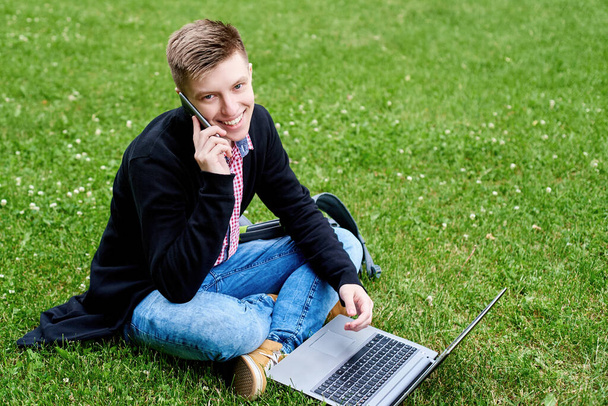 Happy smiling student sitting on green grass lawn, working on laptop computer and talking on cellphone  outdoors, copy space. Preparing for exam, typing, surfing internet. Education concept - Foto, afbeelding