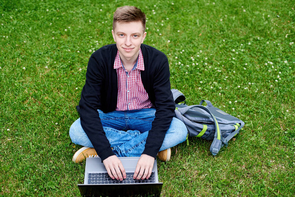 Happy smiling student sitting on green grass lawn in university campus and working on laptop computer outdoors, copy space. Preparing for exam, typing, surfing internet. Education concept - Foto, imagen