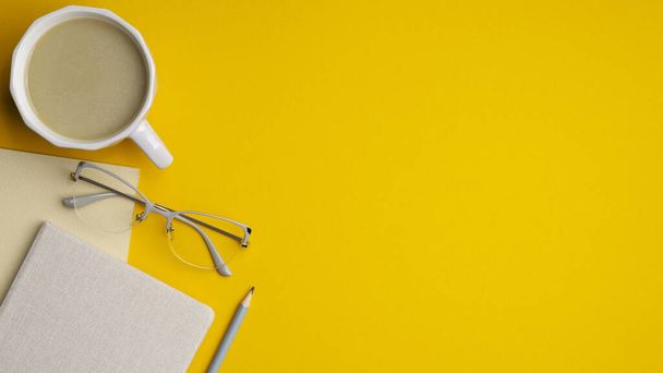 Moderner Schreibtisch mit Tasse Kaffee, Gläsern, Notizbuch und Bleistift auf gelbem Hintergrund. Draufsicht mit Kopierraum, flache Lage. Minimalistisches Arbeitsplatzdesign, stilvoller femininer Arbeitsplatz. - Foto, Bild