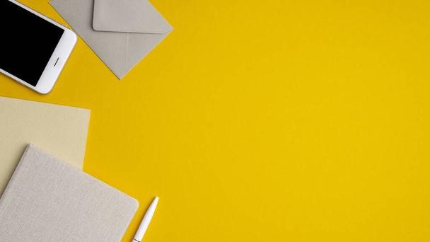 Flat lay, top view office table desk. Workspace with blank notebook, smartphone, envelope, pen on yellow background. Minimal style modern workplace - 写真・画像