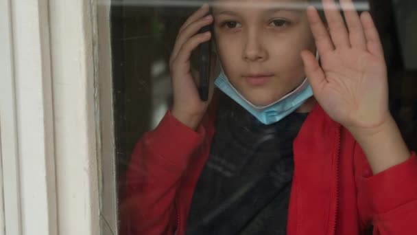 医療マスクの子供は窓の外を見て電話で話す。隔離、コロナウイルス、コビド19における自己分離. - 映像、動画