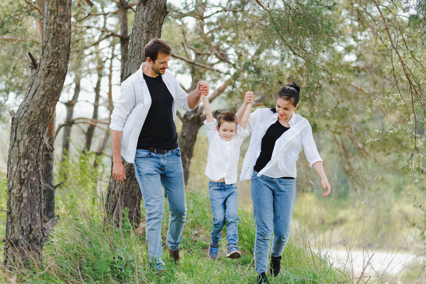 Μαμά, μπαμπάς και γιος περπατάνε στο πράσινο γρασίδι. Χαρούμενη νεαρή οικογένεια που περνάει χρόνο μαζί, τρέχει έξω, πάει στη φύση, σε διακοπές, σε εξωτερικούς χώρους. Η έννοια των οικογενειακών διακοπών. - Φωτογραφία, εικόνα