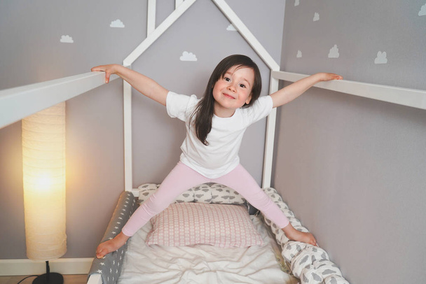 Il bambino sta saltando sul suo letto prima di andare a letto. Una ragazza gioiosa indulge nel suo letto. Bambino in t-shirt bianca e leggings rosa
. - Foto, immagini