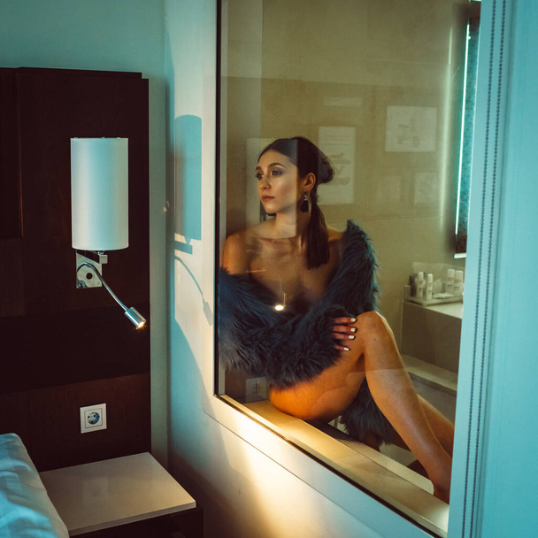 Young beautiful tall girl model half-naked in a blue short coat and black lingerie in a hotel bathroom behind glass - Fotoğraf, Görsel