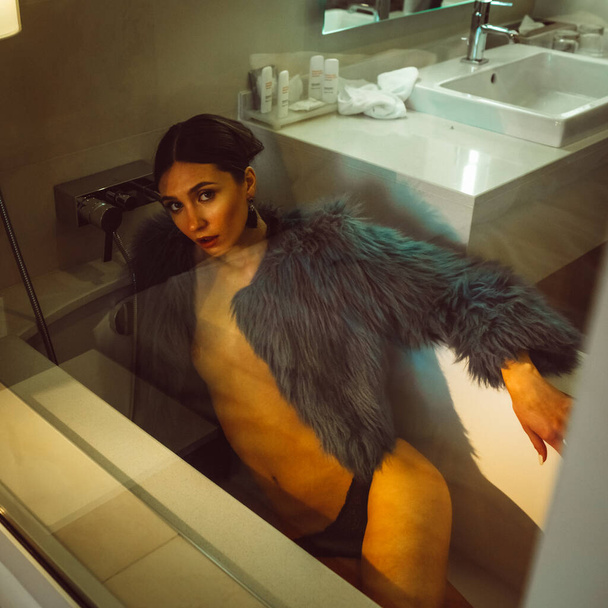 Young beautiful tall girl model half-naked in a blue short coat and black lingerie in a hotel bathroom behind glass - Foto, Imagem