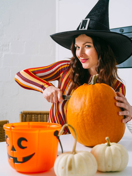 Beautiful young woman wearing witch's black hat  in her kitchen cutting a pumpkin for Halloween. - Zdjęcie, obraz