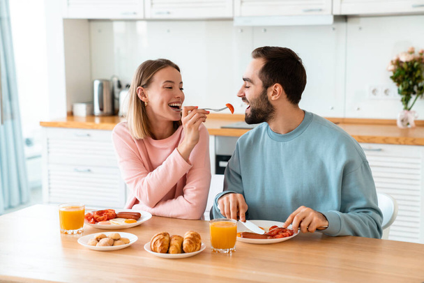 Portrait of young happy couple eating together at table while having breakfast in cozy kitchen at home - Foto, Bild