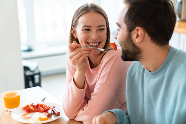 Portrait of young happy couple eating together at table while having breakfast in cozy kitchen at home - Foto, afbeelding