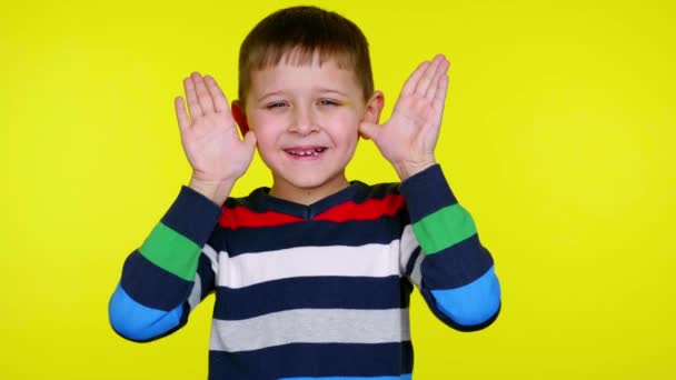 Divertido niño toma las manos de su cara y sonríe en el fondo amarillo
 - Metraje, vídeo