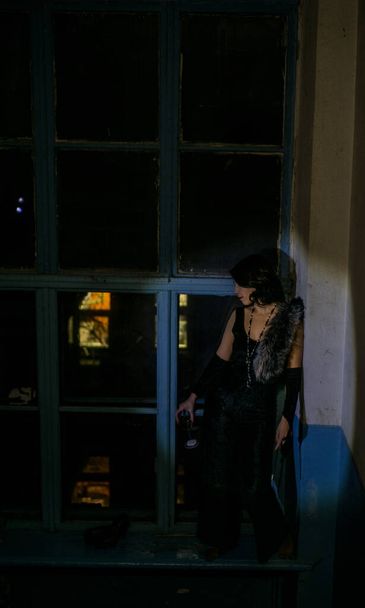 Atrakcyjna kobieta stoi w ciemnym korytarzu naprzeciwko okna, po którym noc lub wieczór. Pani jest ubrana w czarną sukienkę vintage i długie rękawiczki. Patrzy troskliwie na ulicę albo na kogoś.. - Zdjęcie, obraz