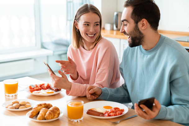 Portrait of young joyful couple using cellphones and laughing while having breakfast in cozy kitchen at home - Zdjęcie, obraz