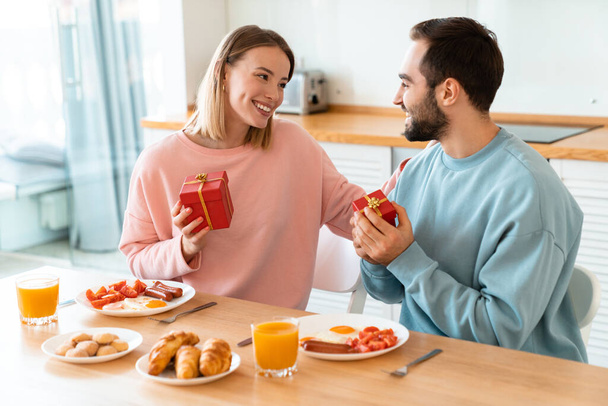 Portret van jong gelukkig paar met geschenkdozen tijdens het ontbijt in gezellige keuken thuis - Foto, afbeelding