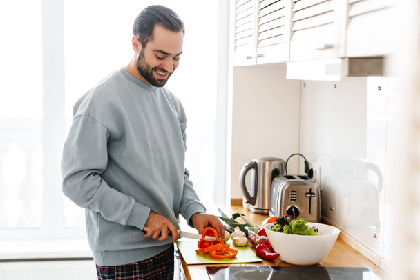 Image of pleased handsome man in casual wear smiling and making lunch in bright modern kitchen - Φωτογραφία, εικόνα