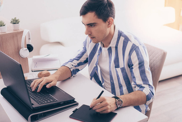 dark-haired man Illustrator in blue shirt draws on graphic tablet, freelancer, remote work on laptop at home, graphic designer concept - Photo, Image
