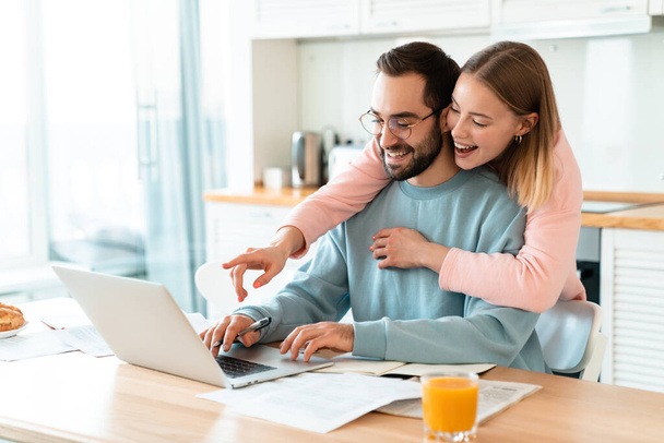 Portrait of young smiling couple hugging while working with laptop and documents in cozy kitchen at home - Фото, изображение