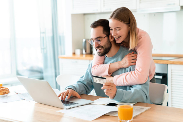Portrait of young joyful couple hugging while working with laptop and holding credit card in cozy kitchen at home - Фото, изображение