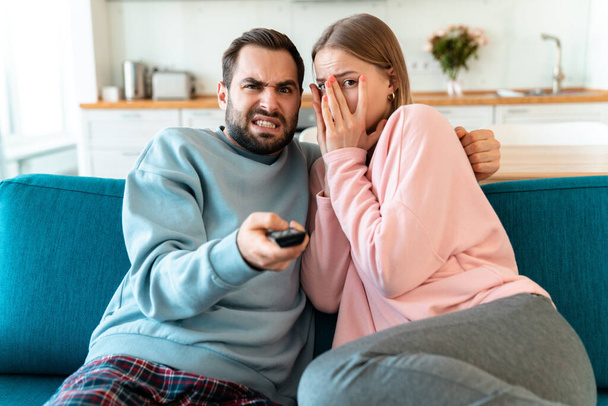 Portrait of young scared couple hugging and watching TV while sitting on sofa in cozy room at home - Foto, afbeelding