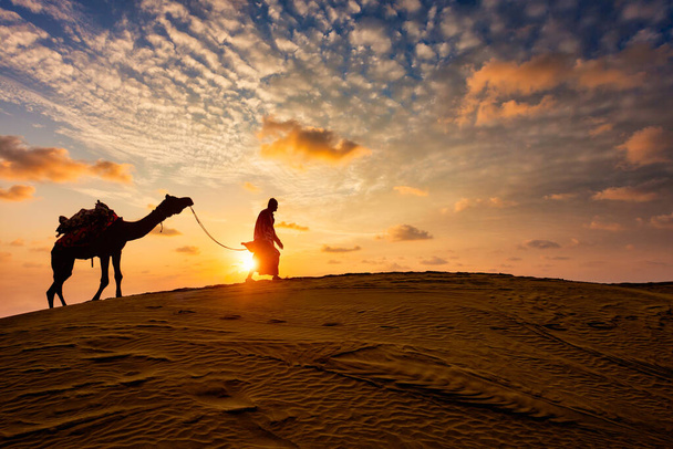 Ινδός οδηγός καμήλας καμήλας με σιλουέτες καμήλας σε αμμόλοφους το ηλιοβασίλεμα. Jaisalmer, Rajasthan, Ινδία - Φωτογραφία, εικόνα