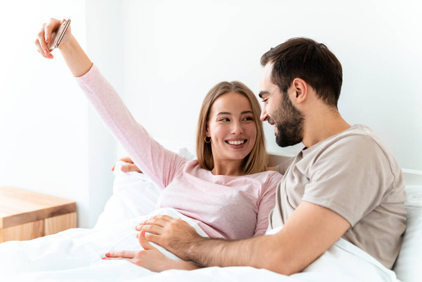 Portrait of beautiful joyful couple hugging and taking selfie photo on smartphone while lying at home bedroom - Foto, afbeelding