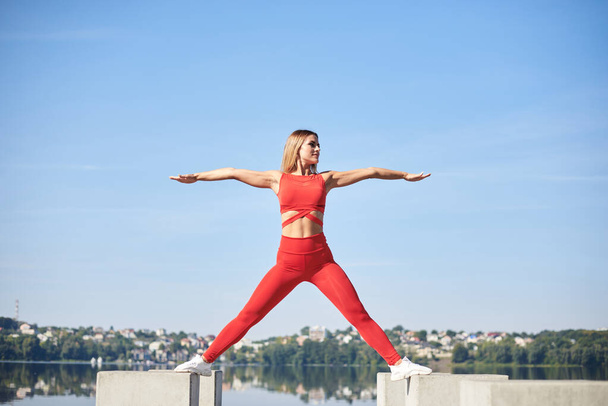Young blond woman, wearing red fitness outfit standing on concrete platform by city lake. Healthy active life concept. Portrait of girl training, doing sports stretching exercise outside in summer. - 写真・画像