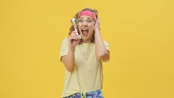 An emotional happy young athletic woman in sportswear is rejoicing while holding a prize isolated over a yellow background in studio - Felvétel, videó